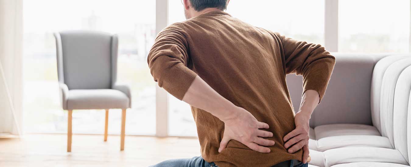 Άνδρας καθισμένος στον καναπέ κρατάει την μέση του, φαίνεται να πονάει.